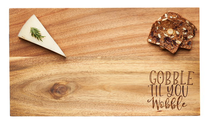 The Maple 14 x 8" Acacia Modern Rectangle Board features "Gobble 'Till You Wobble" engraved in script, shown here with a cheese wedge garnished with rosemary and three seed crackers.