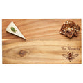 A wedge of herb cheese and seeded crackers sit on Maple’s 14" x 8" Acacia Wood Board, personalized with "The Russells" and a detailed honeybee illustration in the lower right corner.
