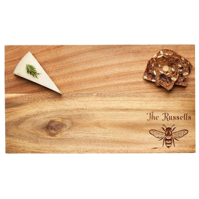 A wedge of herb cheese and seeded crackers sit on Maple’s 14" x 8" Acacia Wood Board, personalized with "The Russells" and a detailed honeybee illustration in the lower right corner.