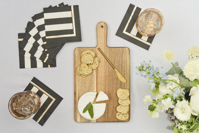 The Sophistiplate 14" x 8" Board Gift Kit: Cordoba holds round brie, crackers, and a gold knife, arranged as an elegant appetizer next to striped napkins, two iced drinks, and a bouquet of white flowers.