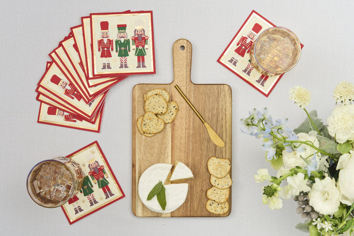 The Sophistiplate 14" x 8" Board Gift Kit: Nutcracker Christmas, featuring a cheese board set with brie, crackers, and a gold knife on acacia wood, sits with festive napkins, drinks, and white flowers on a light tablecloth.