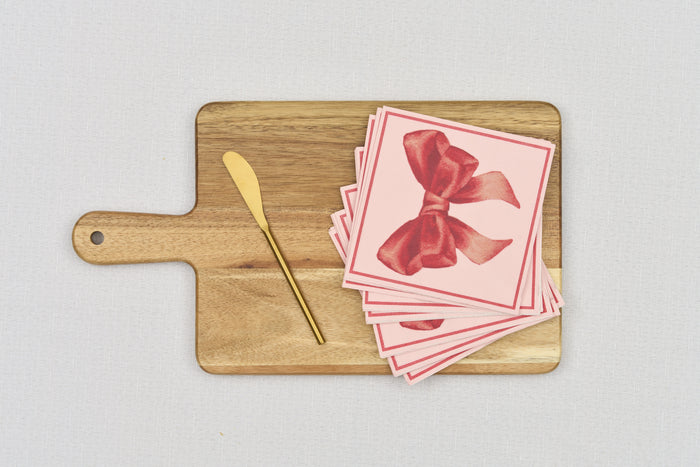 The Sophistiplate 14" x 8" Board Gift Kit includes a wooden cheese board, gold butter knife, and pink napkins with a bold red bow design, arranged on a light surface.