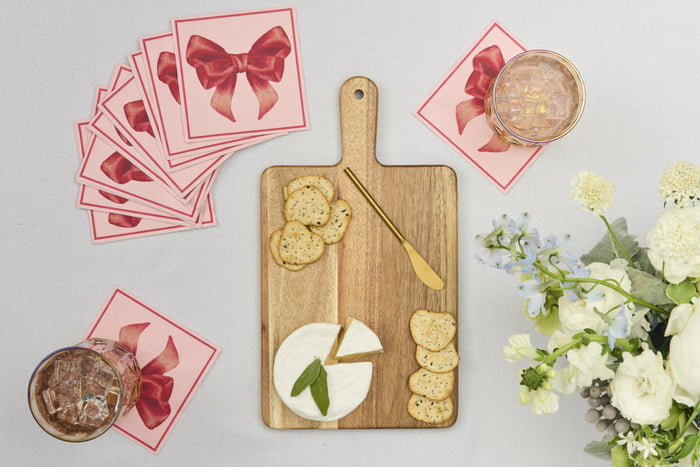 A Sophistiplate 14" x 8" Board Gift Kit features brie, crackers, and a gold knife on acacia wood, with two iced drinks, a floral arrangement, and pink napkins tied with red bows displayed nearby.