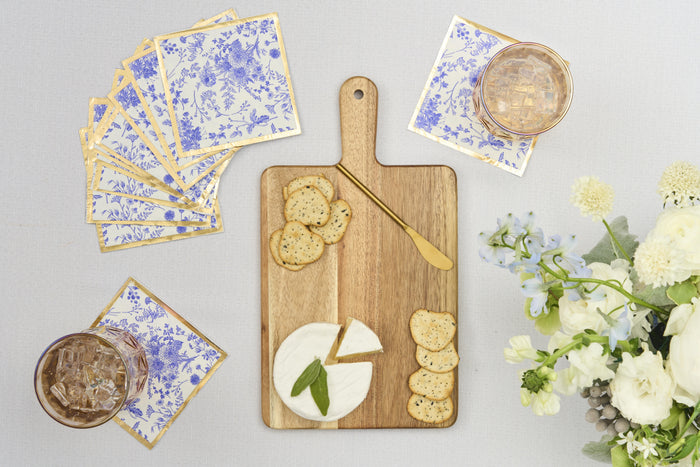 The Sophistiplate 14" x 8" Board Gift Kit: Timeless holds brie, basil leaves, crackers, and a cheese knife on a light surface beside blue-and-white floral napkins, iced drinks, and white blooms—an inviting appetizer tray setup.