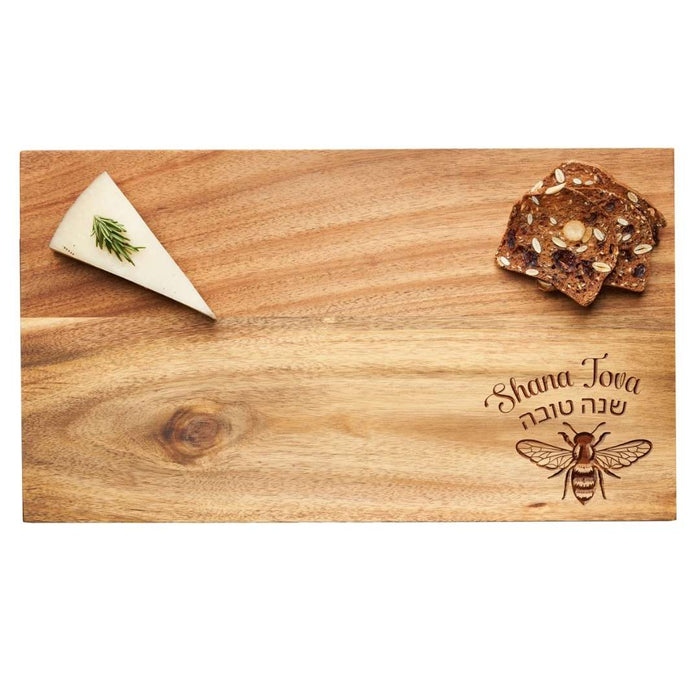 The Maple 14"x8" Acacia Wood Board features "Shana Tova" in English and Hebrew with a bee design, perfect for serving cheese topped with herbs and seedy crackers.
