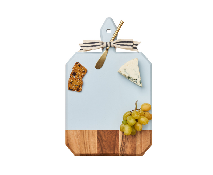 A Maple 16" x 10" Acacia Heirloom Board in Light Blue with a gold spreader displays blue cheese, green grapes, and a fruit and nut cracker, all tied with a striped ribbon, set against a white background.