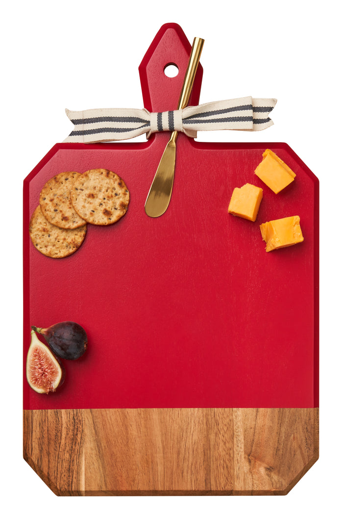 A white background showcases Maple’s 16" x 10" Acacia Heirloom Board in Red with a gold spreader, topped with crackers, cheese cubes, and a halved fig.