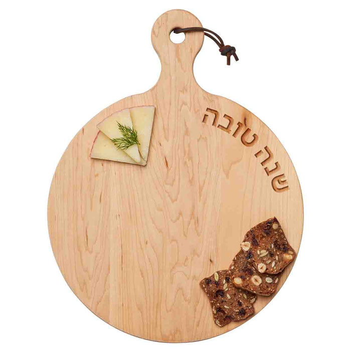 The 16x12" Maple Artisan Round Board - Shana Tova Hebrew Curved by Maple is perfect for Rosh Hashanah, featuring a Hebrew engraving, leather loop handle, and space for crackers, nuts, and cheese slices garnished with dill.