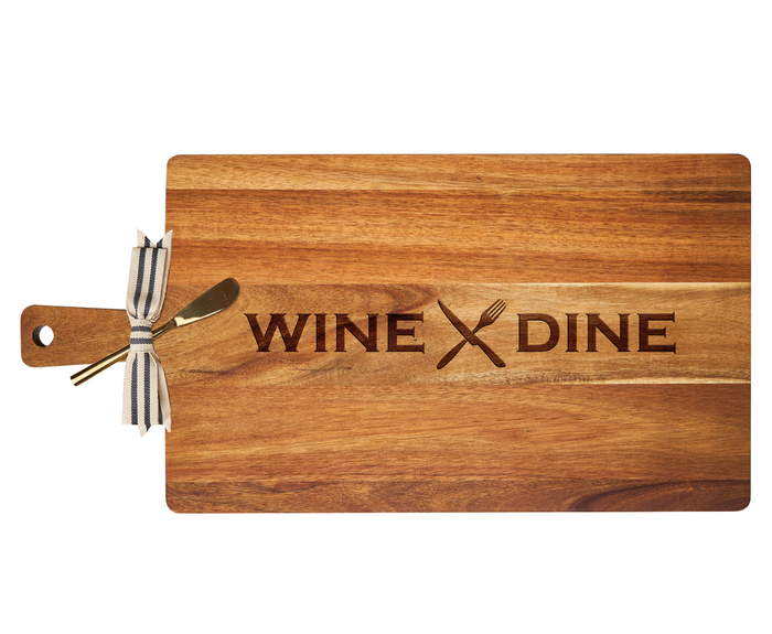 The Maple 24" x 12" Acacia Wood Thick Handle Board features "WINE DINE" and a crossed fork and knife engraving, plus a gold spreader tied with a striped ribbon—combining kitchen decor and functionality.