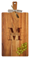The Maple 24" x 12" Acacia Wood Thick Handle Cutting Board features a gold spreader, black-and-white ribbon, and is engraved with a large M and "Morris" in the center, plus includes a bunch of green grapes in the corner.