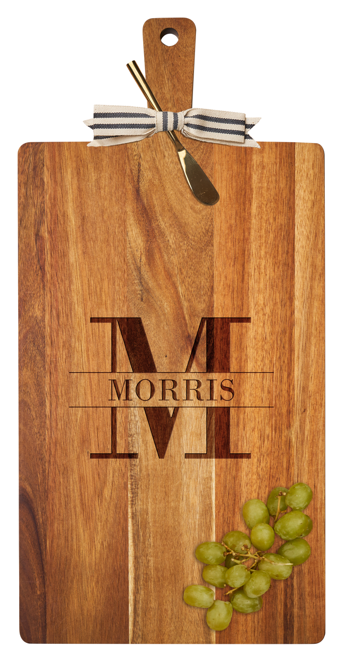 The Maple 24" x 12" Acacia Wood Thick Handle Cutting Board features a gold spreader, black-and-white ribbon, and is engraved with a large M and "Morris" in the center, plus includes a bunch of green grapes in the corner.