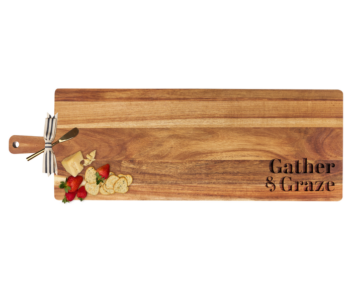 The Maple 35" x 12" Acacia Wood Thick Handle Board features "Gather & Graze" engraving and comes with a gold spreader—ideal for serving cheese, crackers, and fruit while making a charming statement in your kitchen.