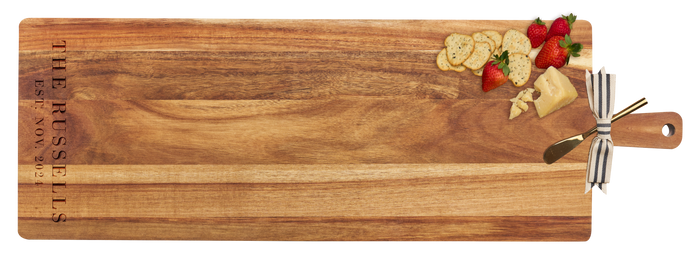 A 35" x 12" Acacia wood Maple charcuterie board with thick handle, engraved with The Russells Est. Nov 2021 on the left, topped with crackers, strawberries, cheese, and a gold spreader tied with a striped ribbon.