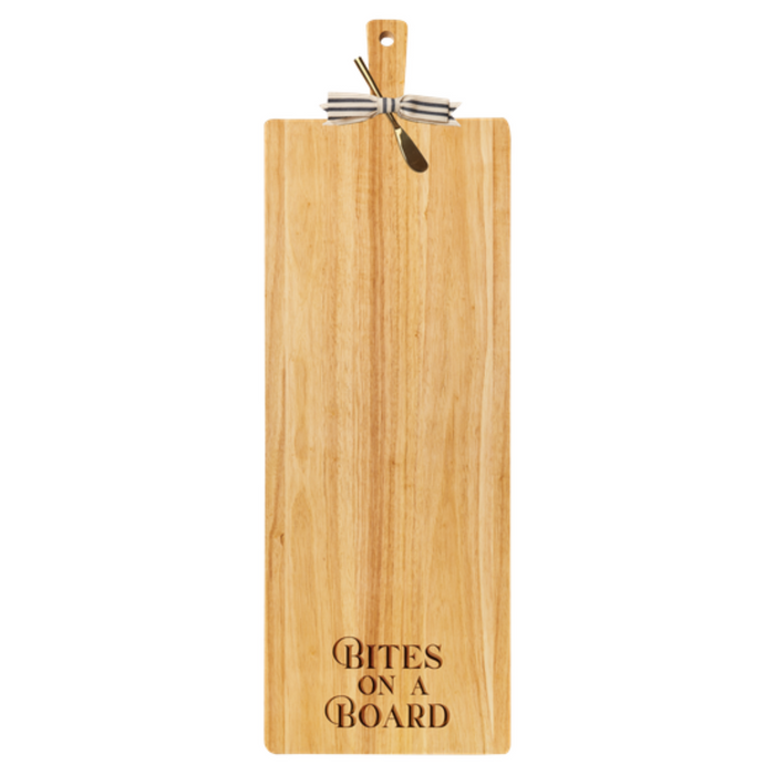 The Maple 35" x 12" Rubber Wood Thick Handle Cutting Board features "BITES ON A BOARD" engraved at the bottom and comes with an elegant metal cheese knife tied to the handle with a black and white ribbon.