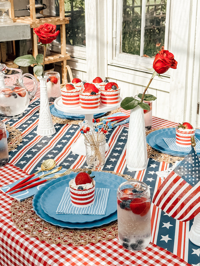 Create a festive table with Sophistiplate LLC's Patriotic French Blue eco-friendly plates, red and white striped desserts with strawberries, berry drinks, red roses in white vases, and American flag decor on a star-and-stripe cloth.