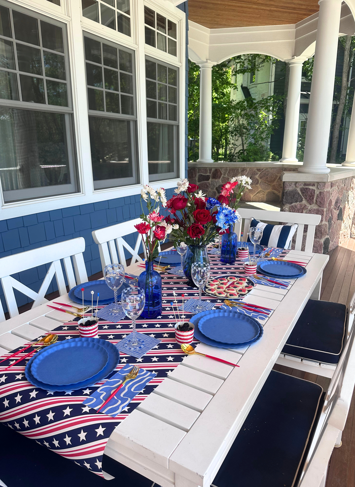 The Sophistiplate LLC 4th of July Table for 16+ in French Blue features eco-friendly disposable plates, red, white, and blue floral arrangements, American flag decor, and a white table with chairs on a porch with columns and large windows.