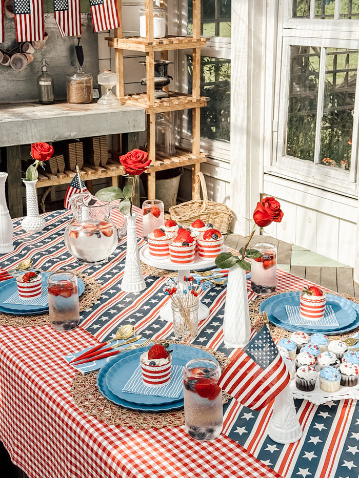 Create a festive patriotic table with Patriotic French Blue by Sophistiplate LLC—red, white, and blue accents, strawberries on desserts, American flags, compostable bamboo plates, red roses in white vases, and a star-striped tablecloth.