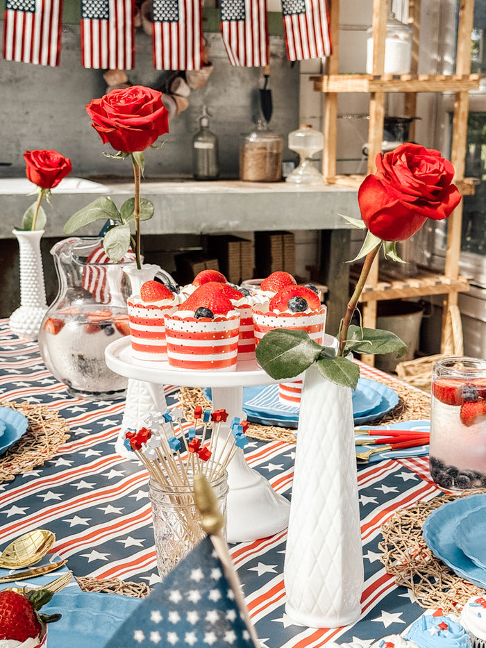 A festive table with red roses, patriotic-themed cupcakes, and American flags showcases Sophistiplate LLC’s Patriotic French Blue collection. Berry desserts in red, white, and blue cups are served on compostable bamboo plates for a sustainable celebration.