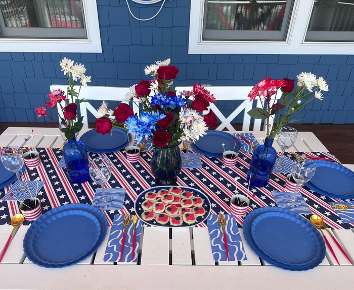 Set a festive scene outdoors with Sophistiplate LLC's 4th of July Table for 16+ in French Blue, featuring eco-friendly plates, star decor, patriotic flowers, and desserts against a blue wall with white windows.