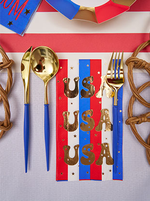 The Modern Patriotic Table Setting by Sophistiplate features gold utensils with blue handles on a red, white, and blue striped napkin with gold USA lettering—perfect festive tableware for parties and celebrations.