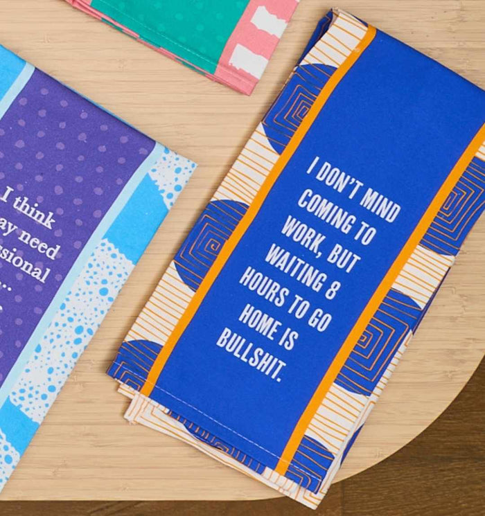The 8 Hours Dish Towel by Drinks On Me is a folded blue towel with orange and white patterns that reads, “I don’t mind coming to work, but waiting 8 hours to go home is bullshit.” Other colorful towels lie nearby on a wooden surface.