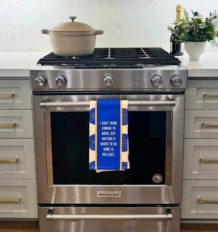 A stainless steel stove holds a beige pot, while the Drinks On Me 8 Hours Dish Towel, featuring a witty saying, hangs from the oven door. A vase with flowers is nearby.