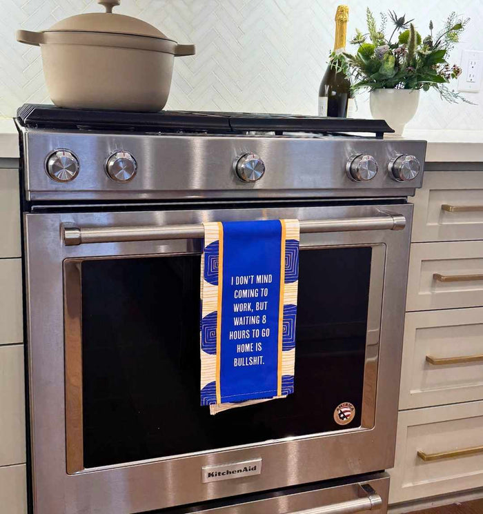 A modern kitchen features a stainless steel oven with the Drinks On Me 8 Hours Dish Towel, blue and yellow, hanging from the door. The towel reads: I DON'T MIND COMING TO WORK, BUT WAITING 8 HOURS TO GO HOME IS BULLSHIT. Pot and flowers sit on the counter.