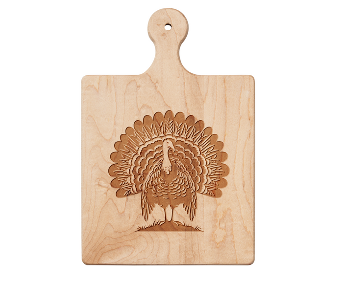 The Maple 9" x 6" Artisan Maple Paddle Board features an engraved turkey with fanned tail feathers on grass and a handle, ideal for use as a charcuterie board.