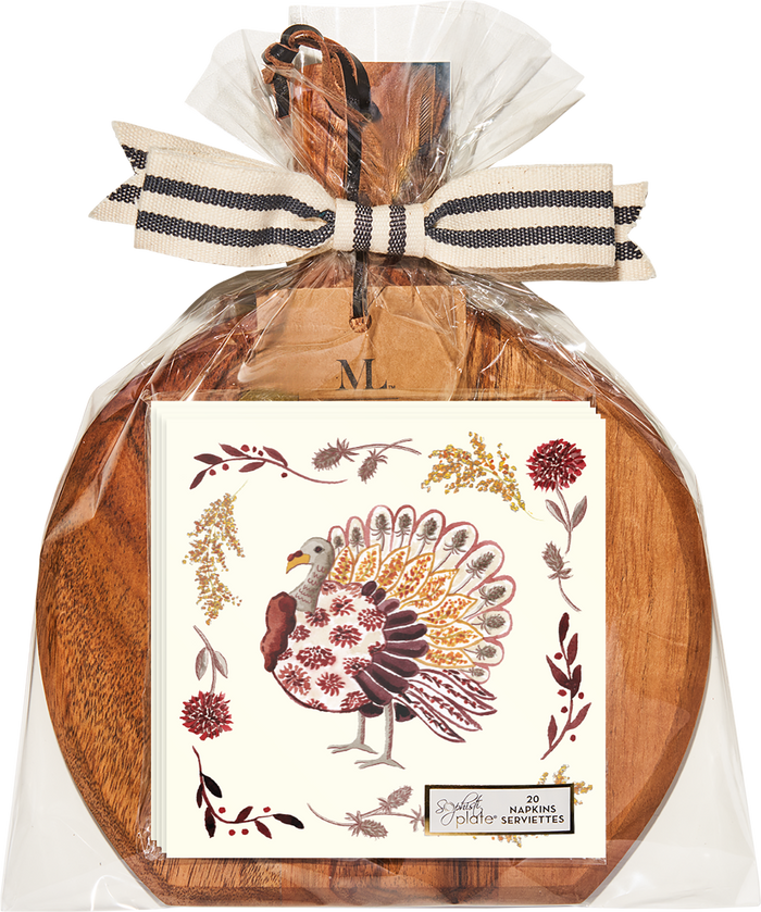 A set of decorative turkey napkins rests elegantly on the Sophistiplate 9.75″ x 8″ Board Gift Kit: Amber Harvest, an acacia wood charcuterie board, wrapped in clear plastic with a striped ribbon and a wooden tag marked M.