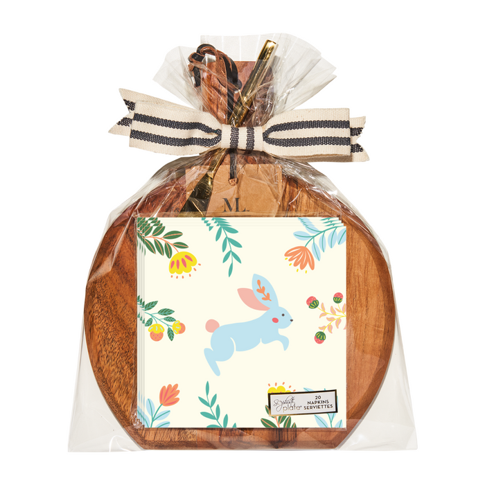 The Sophistiplate 9.75" x 8" Board Gift Kit: Folklore Easter features a wooden board wrapped in clear plastic with a bow, a card showing a blue rabbit with flowers and leaves, kitchen utensils, and a bundle—ideal for charming gifts.