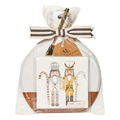 The Maple 9.75" x 8" Board Gift Kit: Gilded Nutcracker includes a round acacia wood board and napkin set with two gold- and silver-accented nutcracker figures, all wrapped in clear plastic with a cream and black striped ribbon.