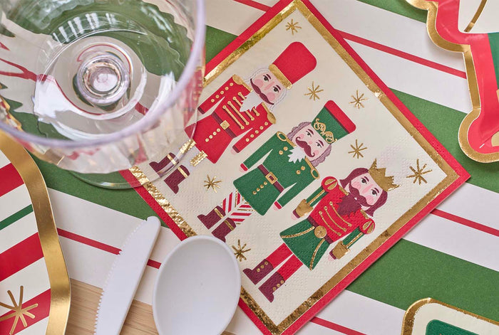 The Nutcracker Christmas Table Setting by Sophistiplate features a festive napkin with three colorful nutcrackers on a red, white, and green striped tablecloth, plus a clear glass, white plastic spoon and knife—all with elegant gold accents.