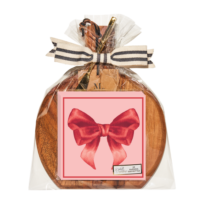 Sophistiplate’s 9.75" x 8" Board Gift Kit: Pink Bows features a wooden serving board and napkins with pink bow designs, wrapped together in clear plastic and finished with a striped ribbon and bow on top.
