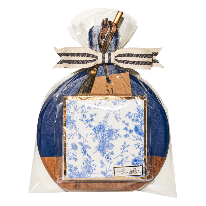 The Maple 9.75" x 8" Board Gift Kit: Timeless is wrapped in clear plastic and features a blue and brown round acacia wood board, white napkins with blue floral designs, a striped bow with a gold pen and tag attached on top.