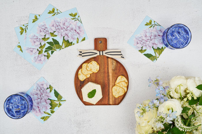 A Sophistiplate 9.75" x 8" Board Gift Kit in Hydrangea sits on a white surface with cheese, crackers, a mint leaf, blue floral napkins, two blue glasses of ice, and a bouquet of white and light blue flowers.