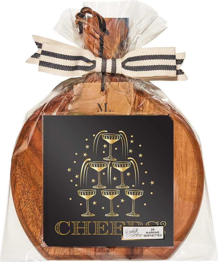 A 9.75″ x 8″ Midnight Sparkle Board Gift Kit by Sophistiplate, beautifully wrapped in clear plastic and tied with a striped ribbon, features a festive card adorned with stacked champagne glasses and the word “Cheers!” in gold. A small wooden tag engraved with the letter “M” is attached to the set, which also includes elegant cocktail napkins for a complete celebratory presentation.