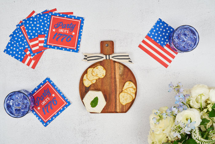 A Sophistiplate 9.75" x 8" Board Gift Kit: Party in the USA displays cheese and crackers on an acacia wood charcuterie board, accented by patriotic cocktail napkins, iced drinks, and a bouquet of white and blue flowers on a white table.
