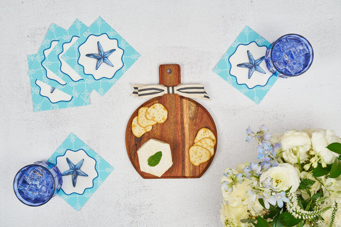 A Sophistiplate 9.75" x 8" Tidal Wave Board Gift Kit displays crackers and cheese atop a white surface, complemented by blue starfish napkins, two blue iced glasses, and a bouquet—an inviting appetizer presentation.