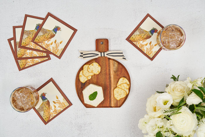 A Sophistiplate 9.75" x 8" Woodland Harvest Board Gift Kit holds cheese and crackers at the center of a white table, surrounded by drinks with ice, four pheasant-themed napkins, and a bouquet of white flowers.