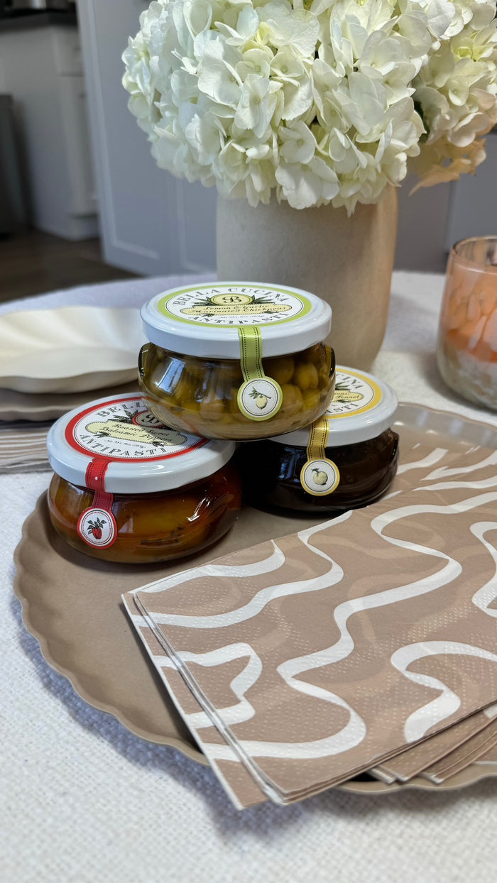 Bella Cucina’s Antipasti Gift Box features three gourmet antipasti jars with colorful seals, stacked on a wavy-edged plate beside patterned napkins—perfect for upgrading your cheese board. White flowers and a candle decorate the table in the background.