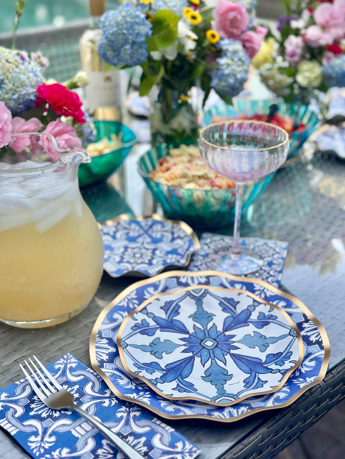 A close-up of a meal set with Sophistiplate LLC’s Moroccan Table Setting, featuring ornate blue and white heavy duty paper plates, a glass of pale drink, napkin, fresh flowers in the background, and a pitcher of iced lemonade nearby.
