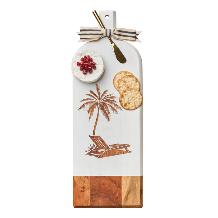 The Maple Acacia Bevel Board Long White w/ Gold Spreader & Ribbon | Palm Tree (20 x 7”) displays round cheese, pomegranate seeds, two crackers, and a gold cheese knife, with a ribbon tied to the handle.