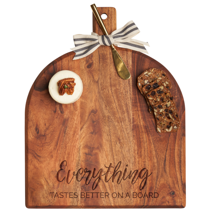 The Maple Acacia Bevel Board (15x12"), engraved with "Everything Tastes Better On A Board," includes a cheese wheel, fruit and nut crackers, a gold spreader knife, and a striped ribbon bow on the handle.