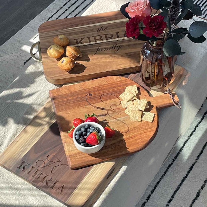 A sunlit table features Maple's 15x12" personalized Acacia Bevel Board with Handle, gold ribbon, and gray/white spreader, displaying pastries, strawberries, crackers, a vase of red flowers, and a bowl of blueberries and blackberries.