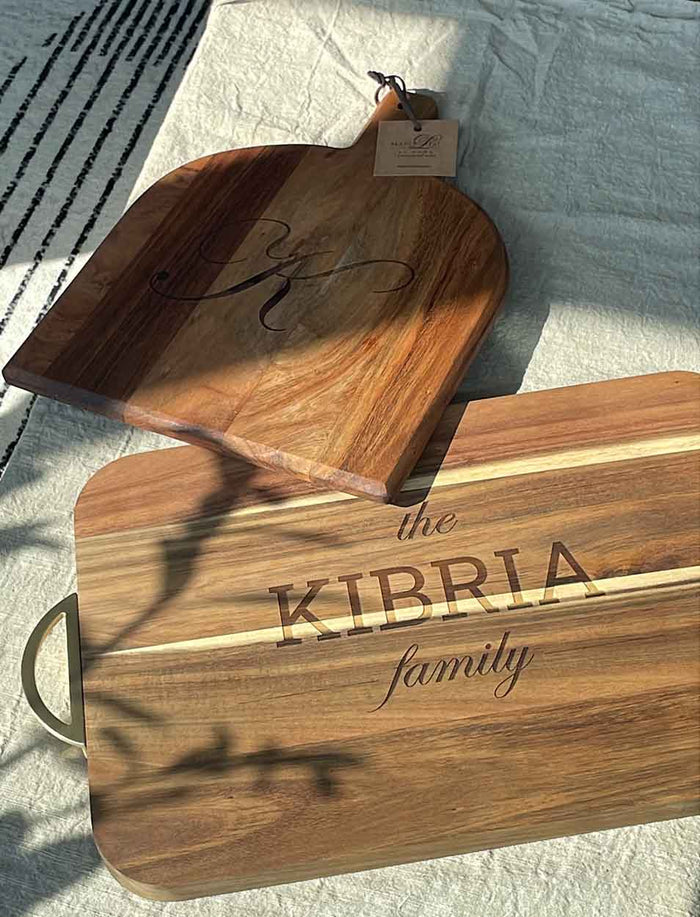 Two Maple Acacia Bevel Boards with Handles rest on a light textured surface: one personalized with a monogrammed K, and another engraved with the KIBRIA family name—both 15x12", food-safe finished, and include a gold ribbon and gray/white spreader.