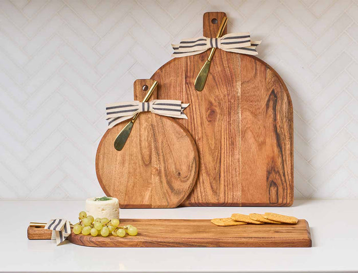 Three Acacia Bevel Boards with Handles by Maple, including a personalized 15"x12" board with a gold ribbon and gray/white spreader, stand upright. The smallest displays cheese, green grapes, and crackers before a white tile wall.