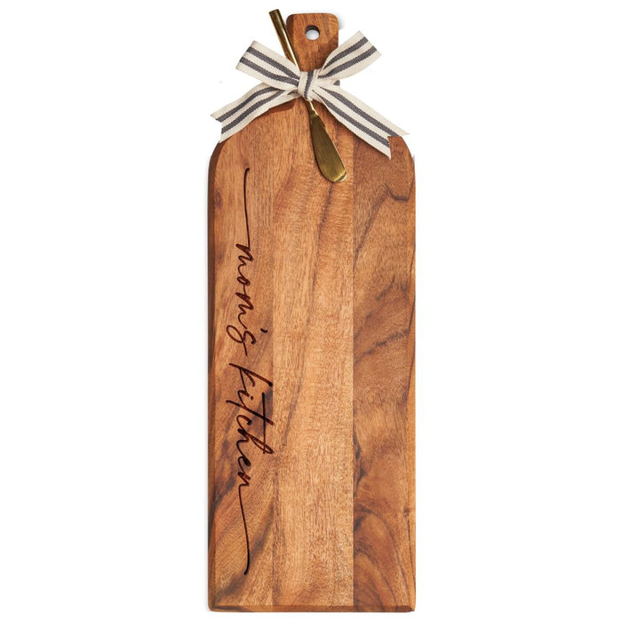The Maple Acacia Bevel Board with Long Handle (20 x 7"), engraved with "Mom's Kitchen" in cursive, includes a striped ribbon accent and a small gold spreader.