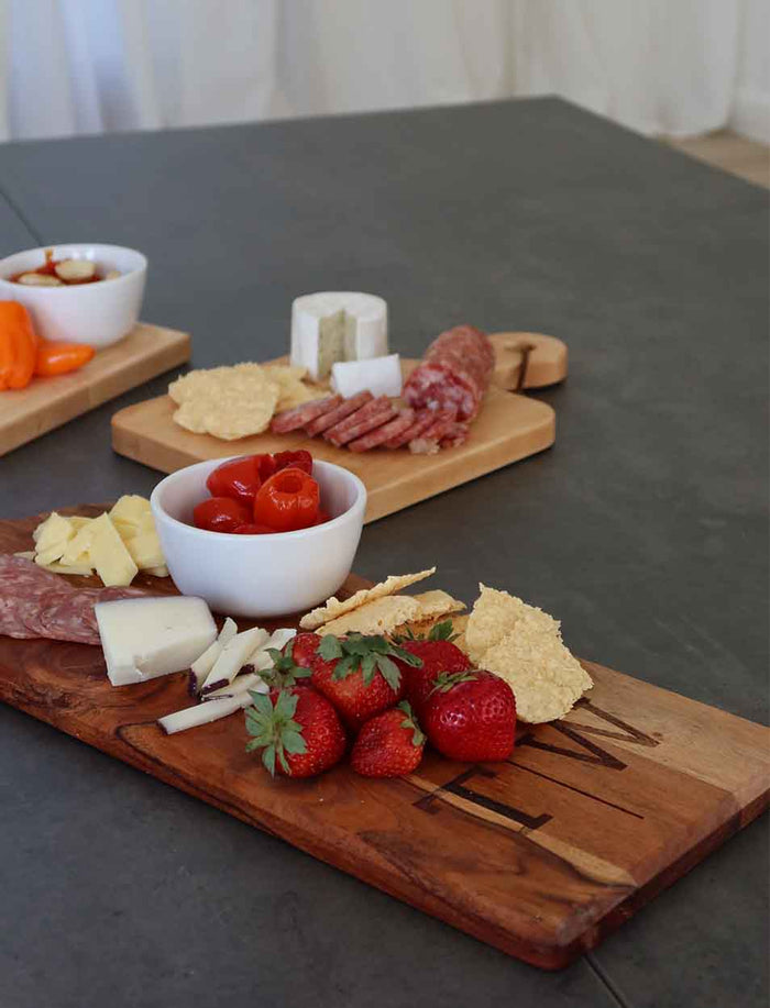 A Maple Acacia Bevel Board with Long Handle (20 x 7", personalized) displays strawberries, cheese, crackers, and salami on a gray table. In the background are two more boards with cheeses, meats, crackers, cherry tomatoes, and dip.