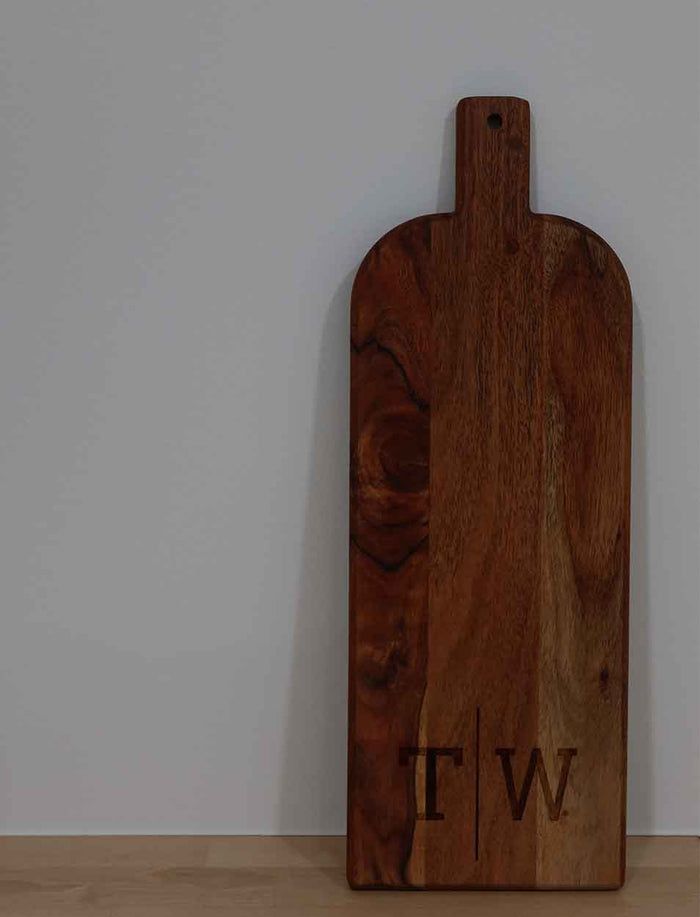 The Maple Acacia Bevel Board with Long Handle (20 x 7") stands upright against a plain white wall, personalized with the engraved monogram "T W" at the bottom right corner.