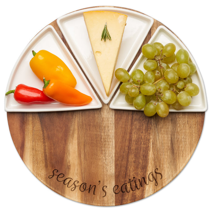 The Maple Acacia Fête Set Round | Season's Eatings | 12" features a 12" acacia wood board engraved with "Season's Eatings," holding three ceramic dishes for serving cheese, mixed bell peppers, and green grapes.